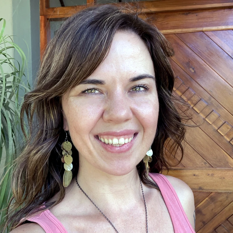 Letitia smiling warmly in natural light, wearing a pink top and shell earrings, standing in front of a wooden door — featured in a testimonial about how Pearl Powder helped heal her eyesight.